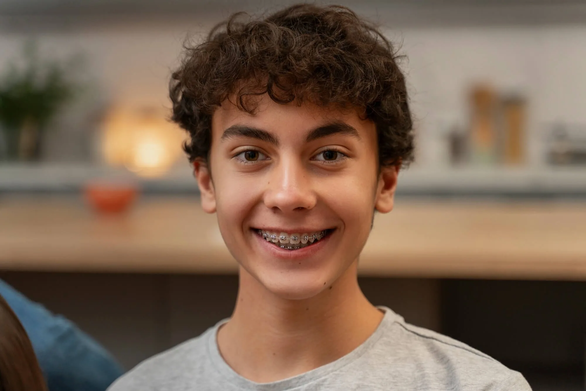 A young boy smiling, showing braces on his teeth, with curly brown hair and wearing a grey t-shirt, indoors with blurred background.